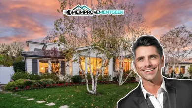 John Stamos House with the actor smiling in front of his charming, warmly lit white cottage surrounded by trees and a lush garden at sunset.