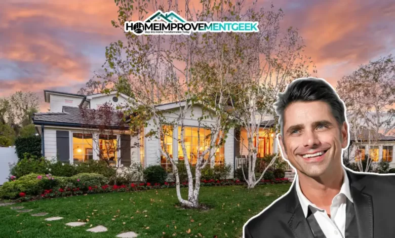 John Stamos House with the actor smiling in front of his charming, warmly lit white cottage surrounded by trees and a lush garden at sunset.