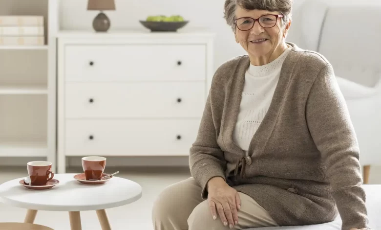 A senior woman sitting on a couch in a cozy aging in place environment, with two cups of coffee on a side table, a dresser with a lamp and books in the background.