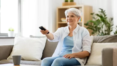 A senior woman comfortably sitting on a couch in a senior-friendly home layout, holding a remote control, with a cup on the table and plants in the background.