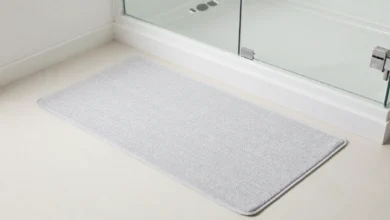 A grey cotton bath mat placed on white bathroom tile outside a glass shower door