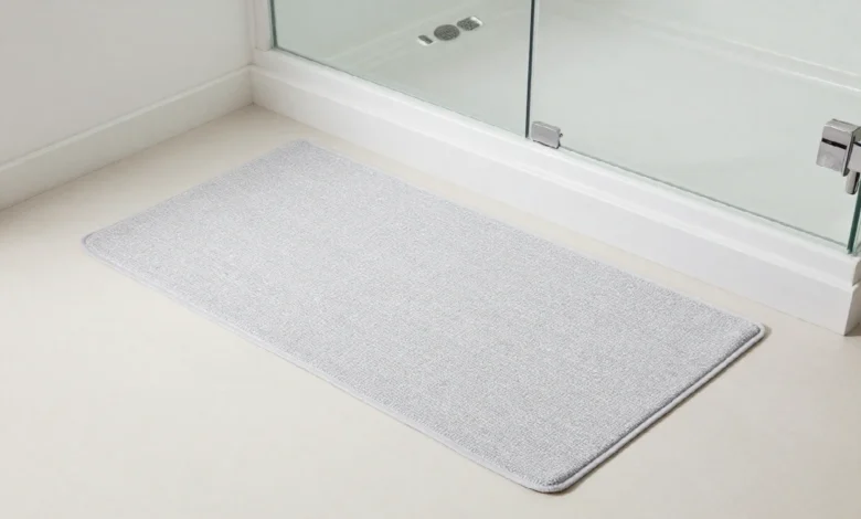 A grey cotton bath mat placed on white bathroom tile outside a glass shower door